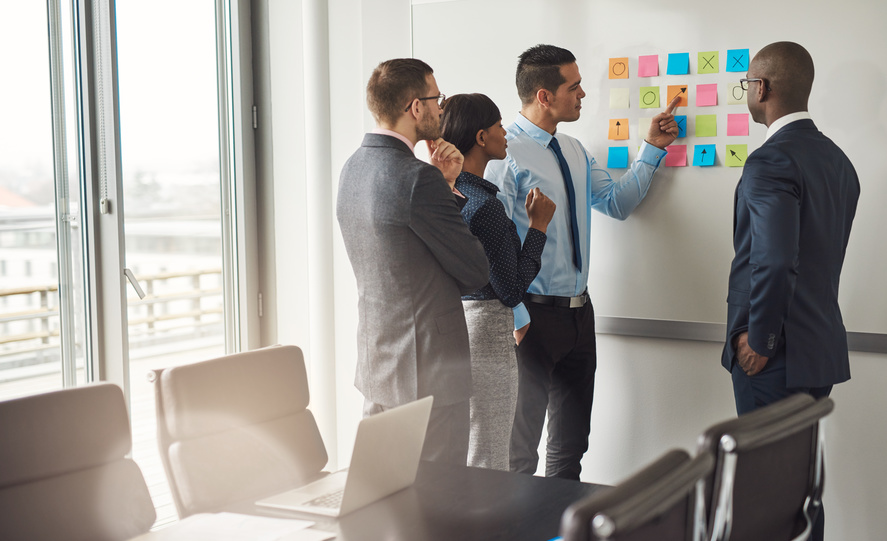 Diverse group of four business people gathered around white board with sticky notes planning something