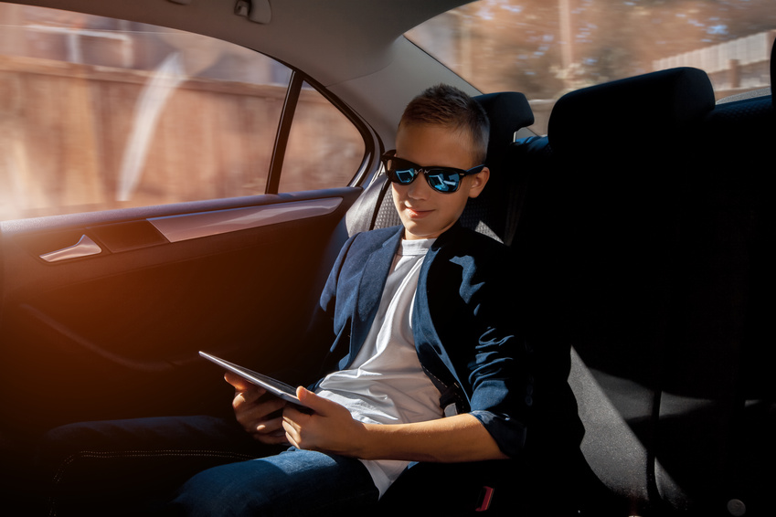 cheerful young boy smiling on a backseat of a car with tablet in hands